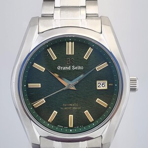 May include: A stainless steel Grand Seiko automatic wristwatch with a green dial and gold-tone markers. The watch features a date window at the 3 o'clock position and a silver bracelet. The watch is a luxury item.
