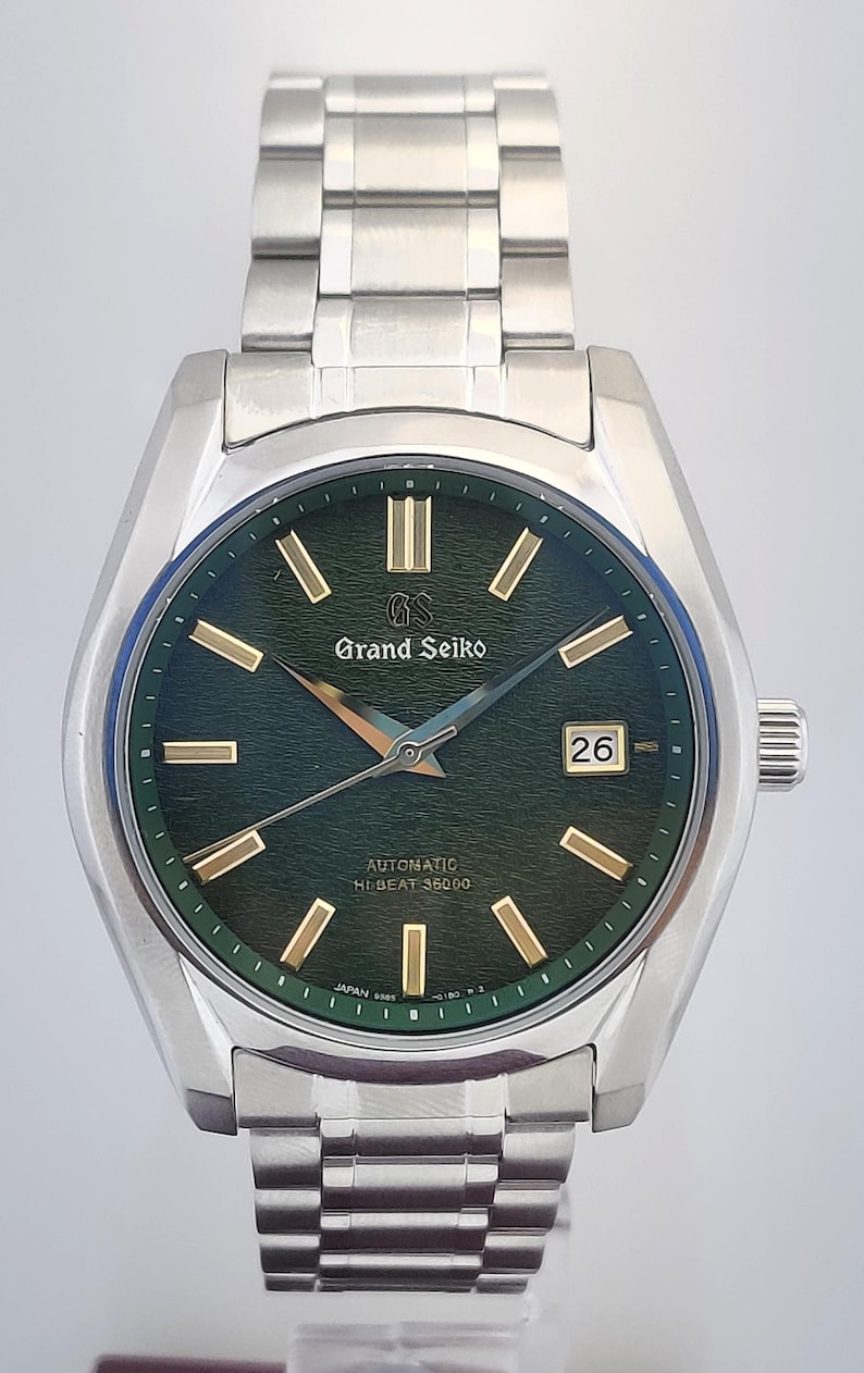 May include: A stainless steel Grand Seiko automatic wristwatch with a green dial and gold-tone markers. The watch features a date window at the 3 o'clock position and a silver bracelet. The watch is a luxury item.