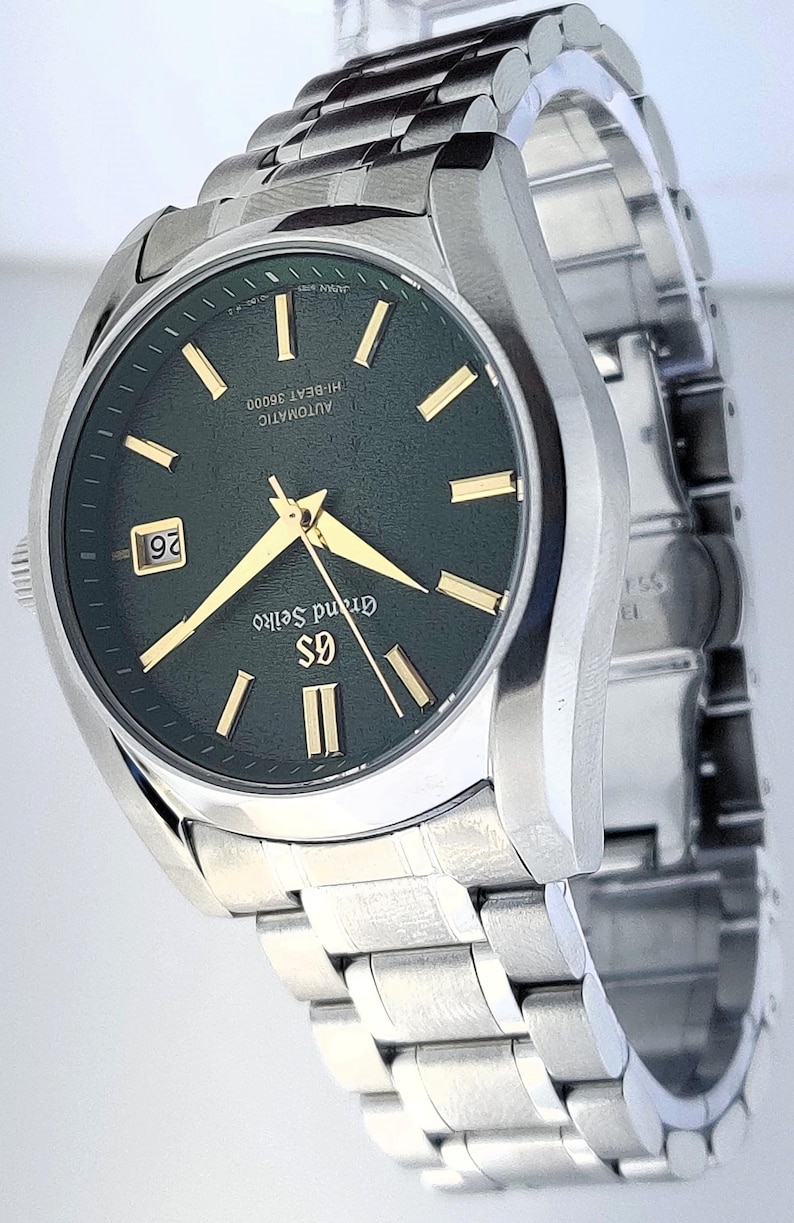 May include: A stainless steel wristwatch with a dark green face and gold-colored hands and markers. The watch has a date display at the 3 o'clock position and a silver-colored band. The watch face has the text "Grand Seiko" and "AUTOMATIC HI-BEAT 36000".