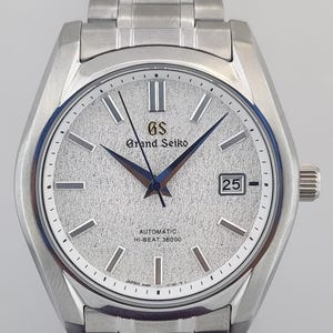 Op de afbeelding: Een zilverkleurig Grand Seiko automatisch polshorloge met een getextureerde zilveren wijzerplaat en een datumvenster op 3 uur. Het horloge heeft blauwe wijzers en uurmarkeringen en een roestvrijstalen armband. De wijzerplaat toont de tekst "Grand Seiko", "Automatic" en "Hi-Beat 36000".