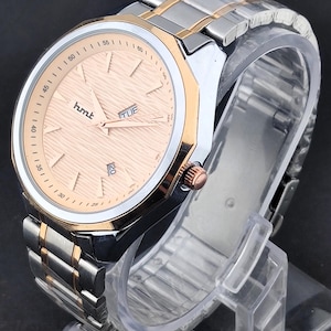 Op de afbeelding: Een roestvrijstalen horloge met goudkleurige accenten. De wijzerplaat is licht perzik van kleur en toont de tekst "hmt" en "TUE". De horlogeband combineert zilveren en goudkleurige schakels. Het horloge wordt gepresenteerd op een transparante plastic standaard.