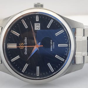 K&ouml;nnte beinhalten: Eine Grand Seiko-Uhr aus Edelstahl mit dunkelblauem Zifferblatt. Die Uhr verf&uuml;gt &uuml;ber silberne Stundenmarkierungen, ein Datumsfenster und ein silbernes Armband. Auf dem Zifferblatt steht "Grand Seiko" und "Spring Drive 5 Days".