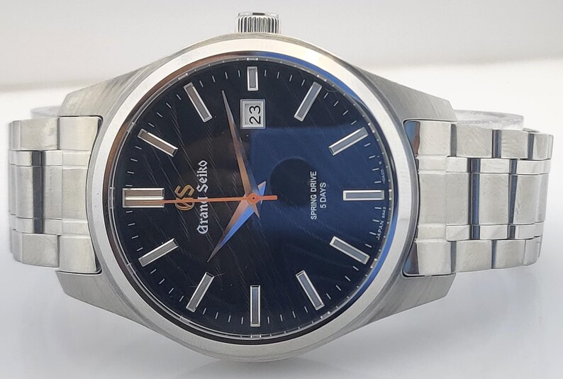K&ouml;nnte beinhalten: Eine Grand Seiko-Uhr aus Edelstahl mit dunkelblauem Zifferblatt. Die Uhr verf&uuml;gt &uuml;ber silberne Stundenmarkierungen, ein Datumsfenster und ein silbernes Armband. Auf dem Zifferblatt steht "Grand Seiko" und "Spring Drive 5 Days".