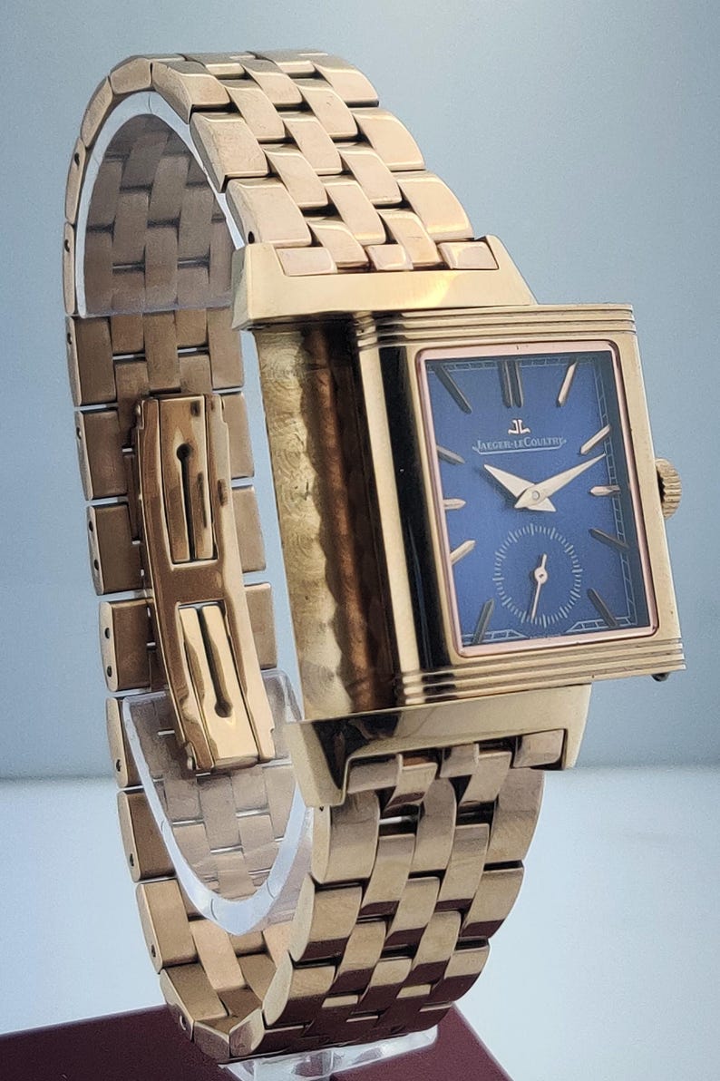 May include: A gold-toned wristwatch with a rectangular blue face and gold hands. The watch features a gold band with a woven design. The watch face has the text "JAEGER-LECOULTRE".