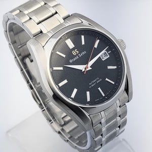 May include: A stainless steel Grand Seiko automatic wristwatch with a black dial. The watch features a date window, silver hands, and a silver bracelet. The dial has the text "Grand Seiko" and "Automatic Hi-Beat 36000".