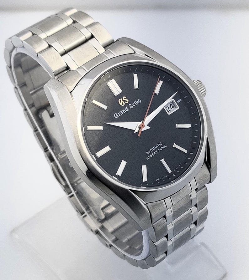 May include: A stainless steel Grand Seiko automatic wristwatch with a black dial. The watch features a date window, silver hands, and a silver bracelet. The dial has the text "Grand Seiko" and "Automatic Hi-Beat 36000".