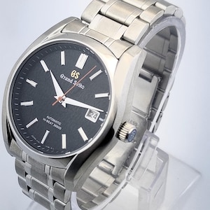 May include: A stainless steel Grand Seiko automatic wristwatch with a black dial. The watch features silver hands and markers, a date window at the 3 o'clock position, and a stainless steel bracelet. The watch is displayed on a clear stand.