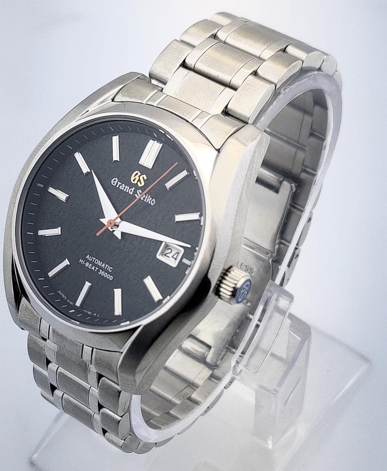May include: A stainless steel Grand Seiko automatic wristwatch with a black dial. The watch features silver hands and markers, a date window at the 3 o'clock position, and a stainless steel bracelet. The watch is displayed on a clear stand.