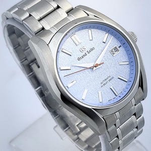 May include: A stainless steel Grand Seiko automatic wristwatch with a light blue textured dial. The watch features a date display and a silver-tone bracelet. The dial includes the text "Grand Seiko" and "AUTOMATIC HI-BEAT 36000".