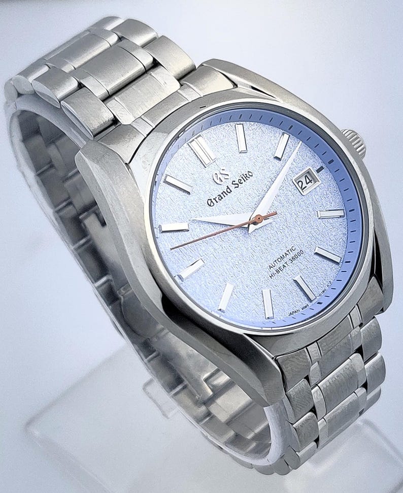 May include: A stainless steel Grand Seiko automatic wristwatch with a light blue textured dial. The watch features a date display and a silver-tone bracelet. The dial includes the text "Grand Seiko" and "AUTOMATIC HI-BEAT 36000".
