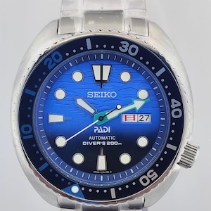 Op de afbeelding: Een roestvrijstalen Seiko PADI automatische duikershorloge met een blauwe wijzerplaat en een zwarte draaibare bezel. De wijzerplaat heeft witte markeringen, een datumweergave en lichtgevende wijzers. De horlogeband is ook van roestvrij staal.
