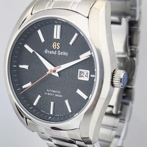 May include: A stainless steel Grand Seiko automatic wristwatch with a dark gray dial. The watch features white hands and markers, a date display, and a silver band. The dial includes the text "Grand Seiko" and "Automatic Hi-Beat 36000".