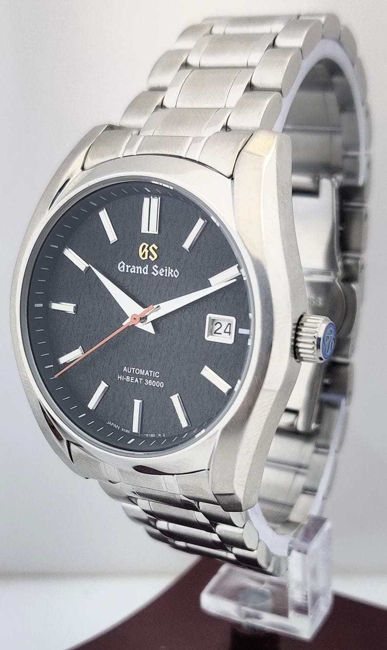 May include: A stainless steel Grand Seiko automatic wristwatch with a dark gray dial. The watch features white hands and markers, a date display, and a silver band. The dial includes the text "Grand Seiko" and "Automatic Hi-Beat 36000".
