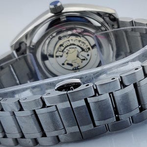 May include: Close-up of a silver wristwatch with a metal band. The watch face is visible, showcasing the intricate inner workings. The watch has a blue crown and a clear protective cover over the band.