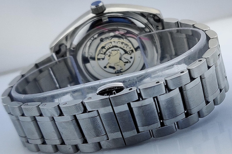 May include: Close-up of a silver wristwatch with a metal band. The watch face is visible, showcasing the intricate inner workings. The watch has a blue crown and a clear protective cover over the band.