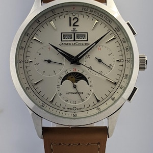 May include: Silver Jaeger-LeCoultre watch with a light brown leather strap. The watch face displays the date, month, and moon phase. The watch has multiple sub-dials and the text "AUTOMATIQUE" and "SWISS MADE".