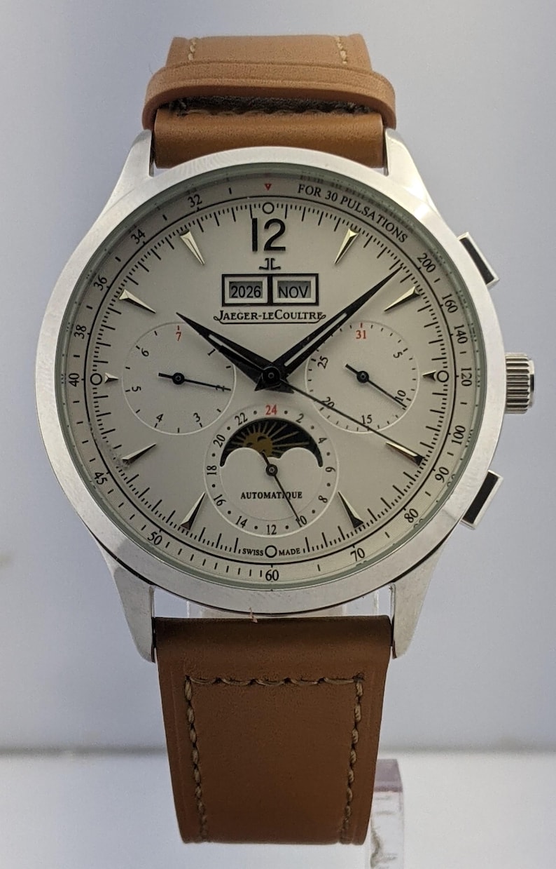 May include: Silver Jaeger-LeCoultre watch with a light brown leather strap. The watch face displays the date, month, and moon phase. The watch has multiple sub-dials and the text "AUTOMATIQUE" and "SWISS MADE".