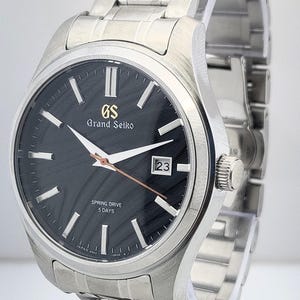 K&ouml;nnte beinhalten: Eine Grand Seiko Armbanduhr aus Edelstahl mit schwarzem Zifferblatt. Die Uhr hat eine Datumsanzeige bei 3 Uhr und ein silbernes Armband. Das Zifferblatt hat silberfarbene Stundenmarkierungen und Zeiger. Eine Luxusuhr.