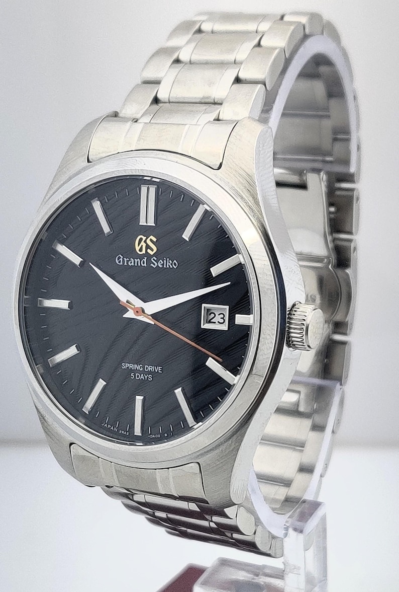 K&ouml;nnte beinhalten: Eine Grand Seiko Armbanduhr aus Edelstahl mit schwarzem Zifferblatt. Die Uhr hat eine Datumsanzeige bei 3 Uhr und ein silbernes Armband. Das Zifferblatt hat silberfarbene Stundenmarkierungen und Zeiger. Eine Luxusuhr.