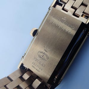 May include: Close-up of a gold-toned Jaeger-LeCoultre Reverso watch. The watch back is visible, with the word "REVERSO" engraved. Additional text includes "3 bar", "1000 hours control", and the numbers "277.8.62" and "2837705".