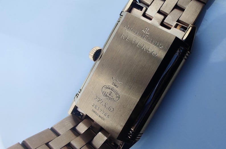May include: Close-up of a gold-toned Jaeger-LeCoultre Reverso watch. The watch back is visible, with the word "REVERSO" engraved. Additional text includes "3 bar", "1000 hours control", and the numbers "277.8.62" and "2837705".