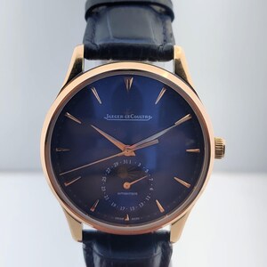 May include: A gold-toned wristwatch with a dark blue face and a black leather band. The watch face features gold-colored hands and markers, with the brand name "Jaeger-LeCoultre" displayed. The watch has a date display and a crown on the side.