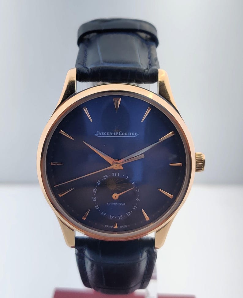May include: A gold-toned wristwatch with a dark blue face and a black leather band. The watch face features gold-colored hands and markers, with the brand name "Jaeger-LeCoultre" displayed. The watch has a date display and a crown on the side.