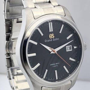 K&ouml;nnte beinhalten: Eine Grand Seiko Spring Drive-Uhr aus Edelstahl mit schwarzem Zifferblatt. Die Uhr verf&uuml;gt &uuml;ber silberne Zeiger und Markierungen, ein Datumsfenster und ein silbernes Armband. Das Zifferblatt zeigt das Grand Seiko-Logo und "Spring Drive 5 Days".
