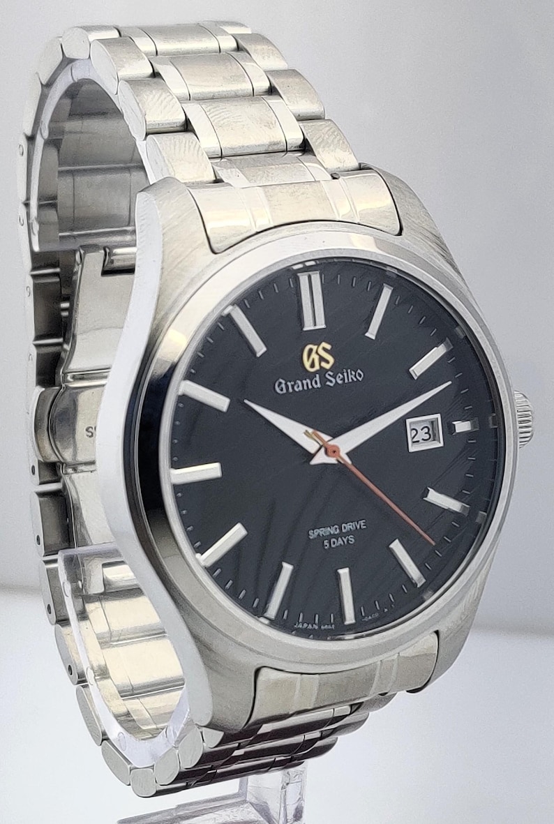 K&ouml;nnte beinhalten: Eine Grand Seiko Spring Drive-Uhr aus Edelstahl mit schwarzem Zifferblatt. Die Uhr verf&uuml;gt &uuml;ber silberne Zeiger und Markierungen, ein Datumsfenster und ein silbernes Armband. Das Zifferblatt zeigt das Grand Seiko-Logo und "Spring Drive 5 Days".