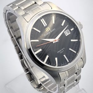 K&ouml;nnte beinhalten: Eine Grand Seiko Spring Drive-Uhr aus Edelstahl mit einem dunkelgrauen Zifferblatt. Die Uhr verf&uuml;gt &uuml;ber silberne Stundenmarkierungen, ein Datumsfenster und ein silbernes Armband. Auf dem Zifferblatt steht "Grand Seiko" und "Spring Drive 5 Days".