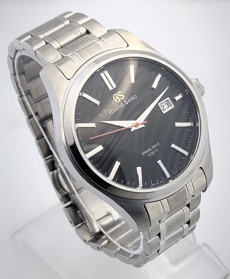 K&ouml;nnte beinhalten: Eine Grand Seiko Spring Drive-Uhr aus Edelstahl mit einem dunkelgrauen Zifferblatt. Die Uhr verf&uuml;gt &uuml;ber silberne Stundenmarkierungen, ein Datumsfenster und ein silbernes Armband. Auf dem Zifferblatt steht "Grand Seiko" und "Spring Drive 5 Days".
