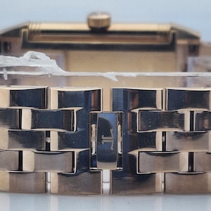 May include: Close-up of a gold-toned wristwatch. The watch features a rectangular face and a band composed of rectangular links. The band is closed with a clasp. The watch is new and still has protective material on it.