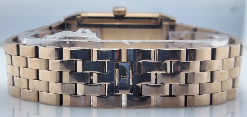 May include: Close-up of a gold-toned wristwatch. The watch features a rectangular face and a band composed of rectangular links. The band is closed with a clasp. The watch is new and still has protective material on it.