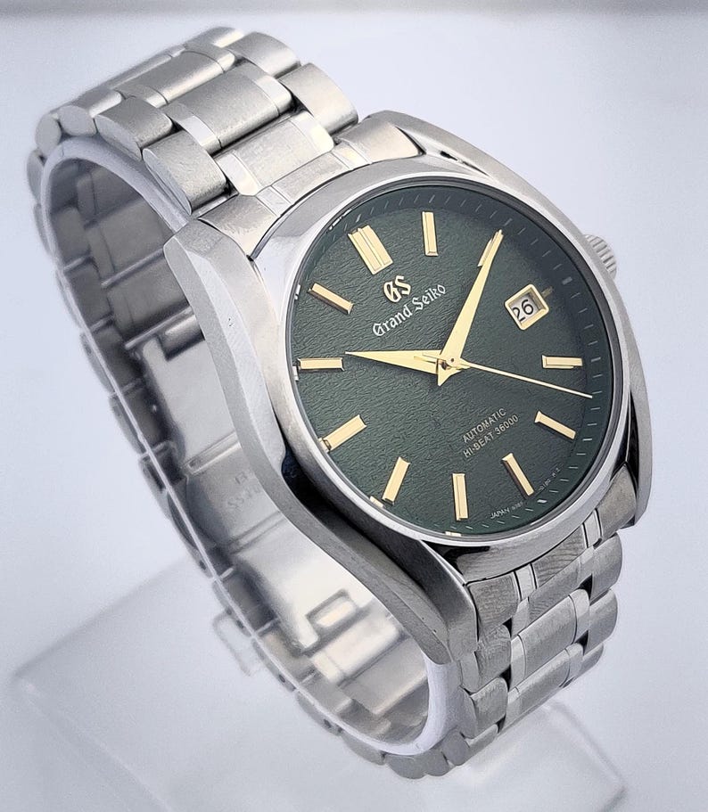 May include: A stainless steel wristwatch with a dark green face and gold-colored hands and markers. The watch has a date display at the 3 o'clock position and a metal band. The watch face has the text "Grand Seiko" and "Automatic Hi-Beat 36000".
