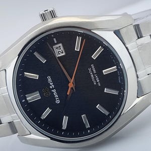 May include: A stainless steel Grand Seiko watch with a black dial. The watch features silver-tone hour markers and hands, a date display, and a metal band. The dial reads "Grand Seiko" and "AUTOMATIC HI-BEAT 36000".