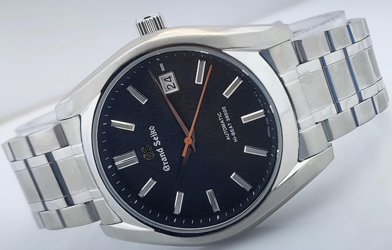May include: A stainless steel Grand Seiko watch with a black dial. The watch features silver-tone hour markers and hands, a date display, and a metal band. The dial reads "Grand Seiko" and "AUTOMATIC HI-BEAT 36000".