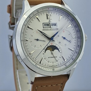 May include: Silver-toned Jaeger-LeCoultre watch with a light brown leather strap. The watch face displays multiple subdials, a moon phase indicator, and the date 2026 NOV. The watch has the text "AUTOMATIQUE" and "SWISS MADE".