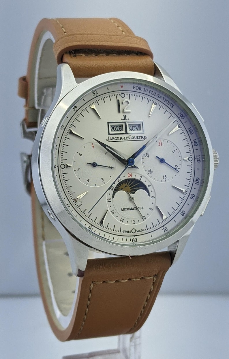 May include: Silver-toned Jaeger-LeCoultre watch with a light brown leather strap. The watch face displays multiple subdials, a moon phase indicator, and the date 2026 NOV. The watch has the text "AUTOMATIQUE" and "SWISS MADE".