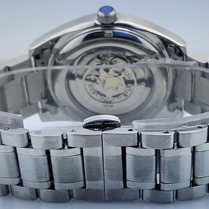 May include: Close-up of a silver-toned wristwatch with a stainless steel band. The watch face is visible through the transparent back, revealing the intricate inner workings. The band is secured with a clasp.