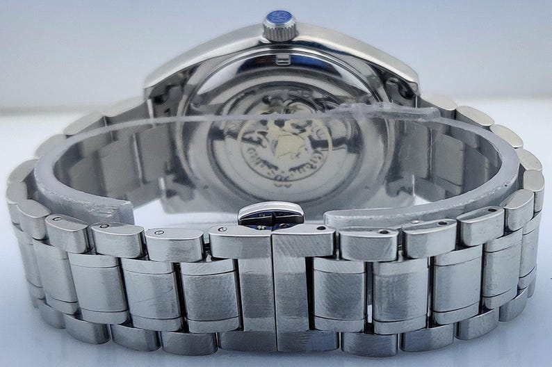 May include: Close-up of a silver-toned wristwatch with a stainless steel band. The watch face is visible through the transparent back, revealing the intricate inner workings. The band is secured with a clasp.