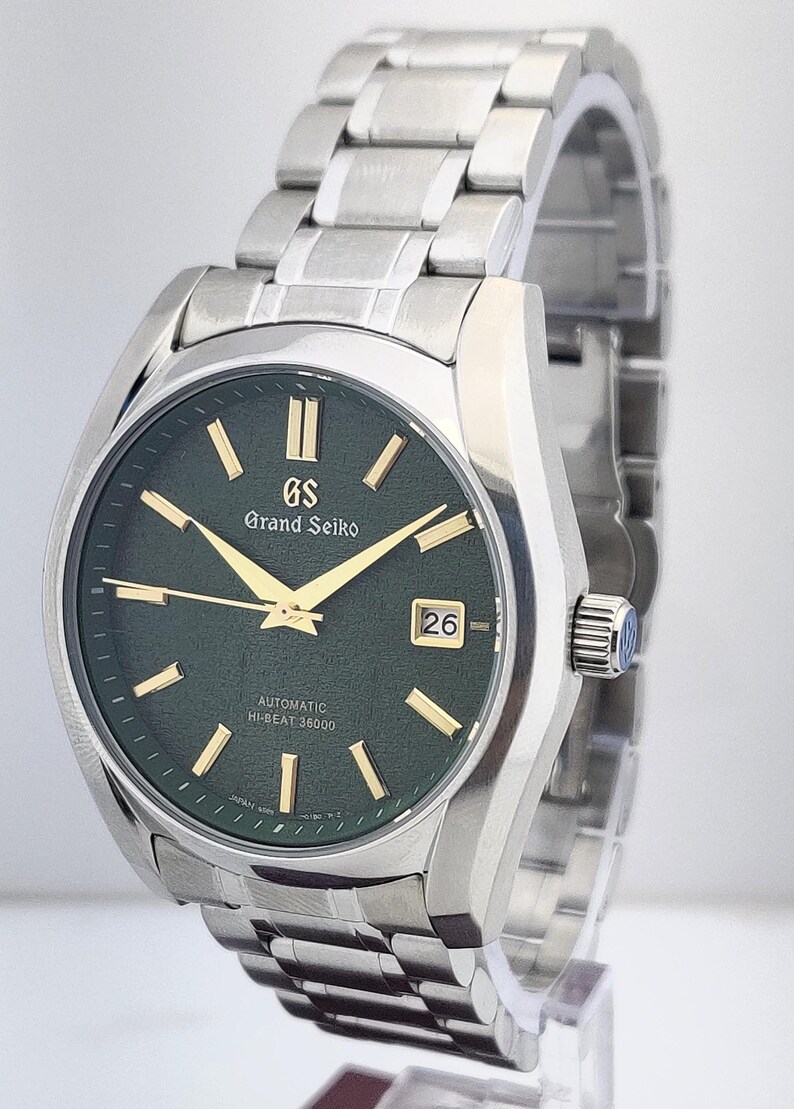 May include: A stainless steel Grand Seiko automatic watch with a dark green dial. The watch features gold-colored hands and markers, a date window at the 3 o'clock position, and the text "Grand Seiko" and "Automatic Hi-Beat 36000".