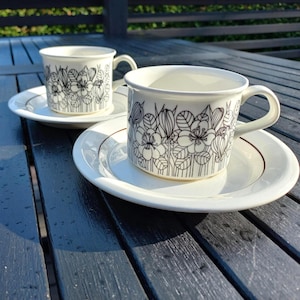 Op de afbeelding: Twee witte keramische koffiekopjes en schotels. Elk kopje en schotel heeft een zwart bloemmotief. De kopjes hebben een oor en staan op een witte schotel met een dunne bruine rand. Op een donker houten oppervlak.
