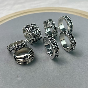 May include: A collection of six silver rings featuring intricate embossed designs, including floral scrollwork, cross motifs, and a sword detail. The rings vary in width and thickness, with some displaying visible hallmarks and engraved text on the inner bands.