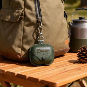 Puede incluir: Una funda de cuero verde oscuro para AirPod con el nombre "Dominic" grabado. La funda está sujeta a una mochila con un mosquetón plateado. Un cono de pino y una tetera de camping están sobre una mesa de madera.
