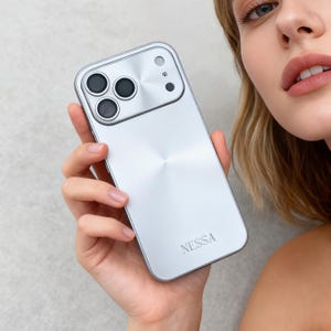 Puede incluir: Una funda plateada para teléfono con un diseño circular alrededor de las lentes de la cámara y la palabra "NESSA" impresa en la parte inferior. La funda tiene un acabado de metal cepillado y se sostiene en una mano.