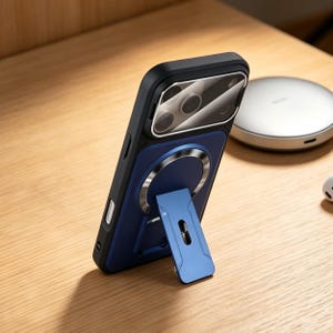 Könnte beinhalten: Eine schwarz-blaue Handyhülle mit einem silbernen Ring und einem blauen Ständer. Die Hülle liegt auf einer Holzoberfläche, im Hintergrund ein kabelloses Ladegerät. Die Handyhülle dient zum Schutz des Telefons und ermöglicht das Aufstellen.