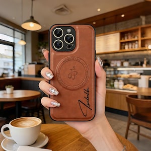 Puede incluir: Una funda de cuero marrón para teléfono con marco de cámara negro y un diseño de anillo magnético circular en la parte posterior. La funda presenta un logo con las letras JJ dentro del círculo magnético y el nombre Isabella en cursiva negra.