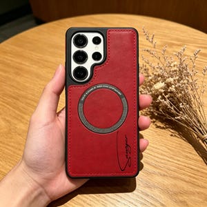 Könnte beinhalten: Rote und schwarze Handyhülle mit einem Magnetring und dem Namen "Sawyer" in Schreibschrift. Die Hülle hat eine strukturierte lederartige Optik und einen schwarzen Rand. Die Kameralinsen des Telefons sind sichtbar.