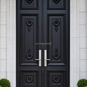 May include: A black double door with decorative floral carvings and silver handles. The door is framed by a white stone facade and includes the text "carvawood". Green bushes in dark grey planters flank the entrance.