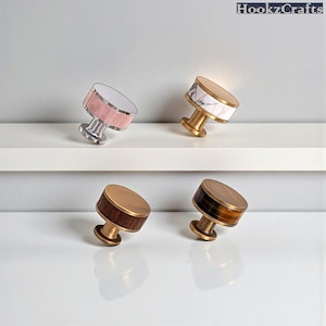 Puede incluir: Cuatro tiradores decorativos para armarios con varios diseños. Uno tiene un diseño rosa y plateado, otro un diseño de mármol blanco y dorado, y los otros dos tienen un diseño de madera y dorado. Los tiradores están colocados sobre una estantería y una superficie blancas.